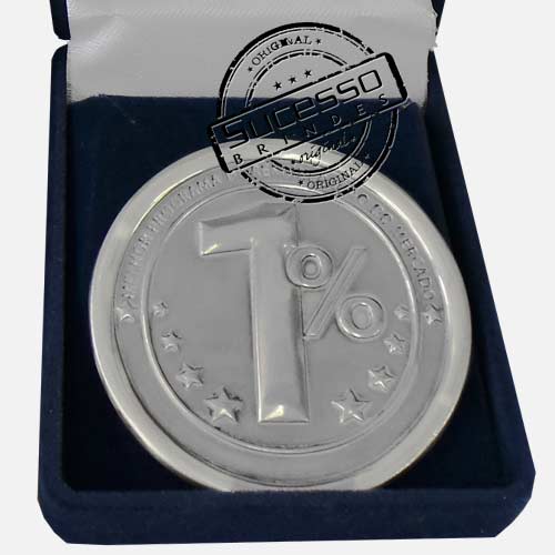 Medalha prateada com o símbolo '1%' gravado no centro, cercada por pequenas estrelas ao redor, dentro de uma caixa de veludo azul. O selo 'Sucesso Brindes Original' está sobreposto à imagem.